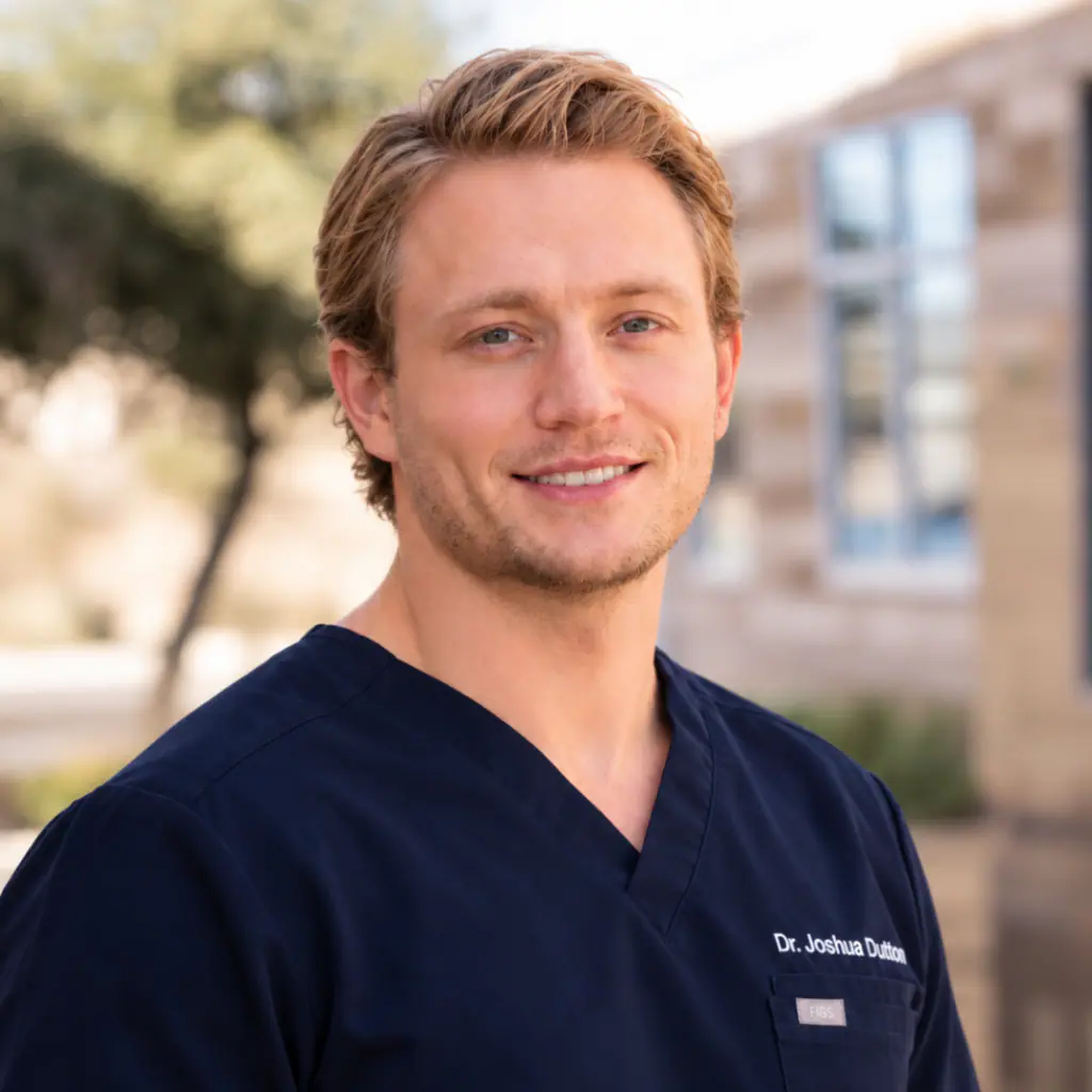 Portrait of a smiling male doctor in navy scrubs standing outdoors, name Dr. Joshua Dutton embroidered on chest.